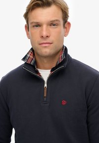 Marineblauwe pullover met een halve rits kraag heeft een vuilnisvoering. Inclusief een rood logo op de borst en een textuur van katoenmix.