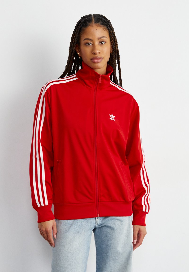 Giacca rossa con zip realizzata in tessuto liscio, con strisce bianche sulle maniche e un piccolo logo sul petto. Include tasche laterali.