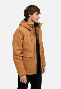 Brun hættejakke med lynlås foran, sidelommer og en afslappet pasform. Lavet af struktureret stof, båret over en sort skjorte og mørke bukser.