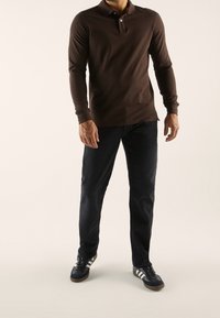 Ruskea pitkähihoinen poolopaita, jossa on kaulus ja kaksi nappia, yhdistetty tummansinisiin farkkuihin ja mustiin, valkoisilla raidoilla varustettuihin urheilukenkiin.