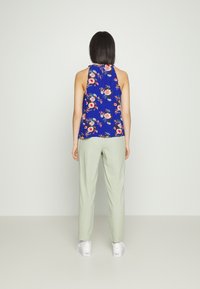 Růžový top bez rukávů s květinovým vzorem a vysokým límcem, doplněný volnými světle zelenými kalhotami a bílými teniskami, z pohledu zezadu.