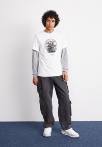 Camiseta blanca con un gráfico de baloncesto, capa base de manga larga gris, pantalones cargo grises y zapatillas blancas. Diseño simple con bolsillos prácticos.