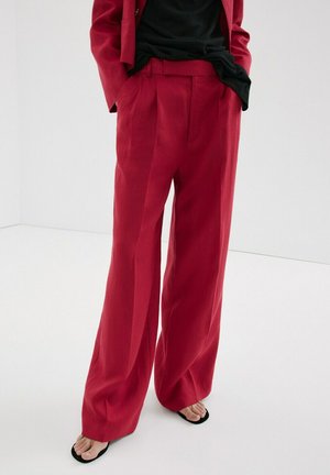 Person trägt hoch taillierte, weit geschnittene rote Hosen mit Händen in den Taschen, kombiniert mit schwarzen Flip-Flops und einem teilweise sichtbaren schwarzen Oberteil.