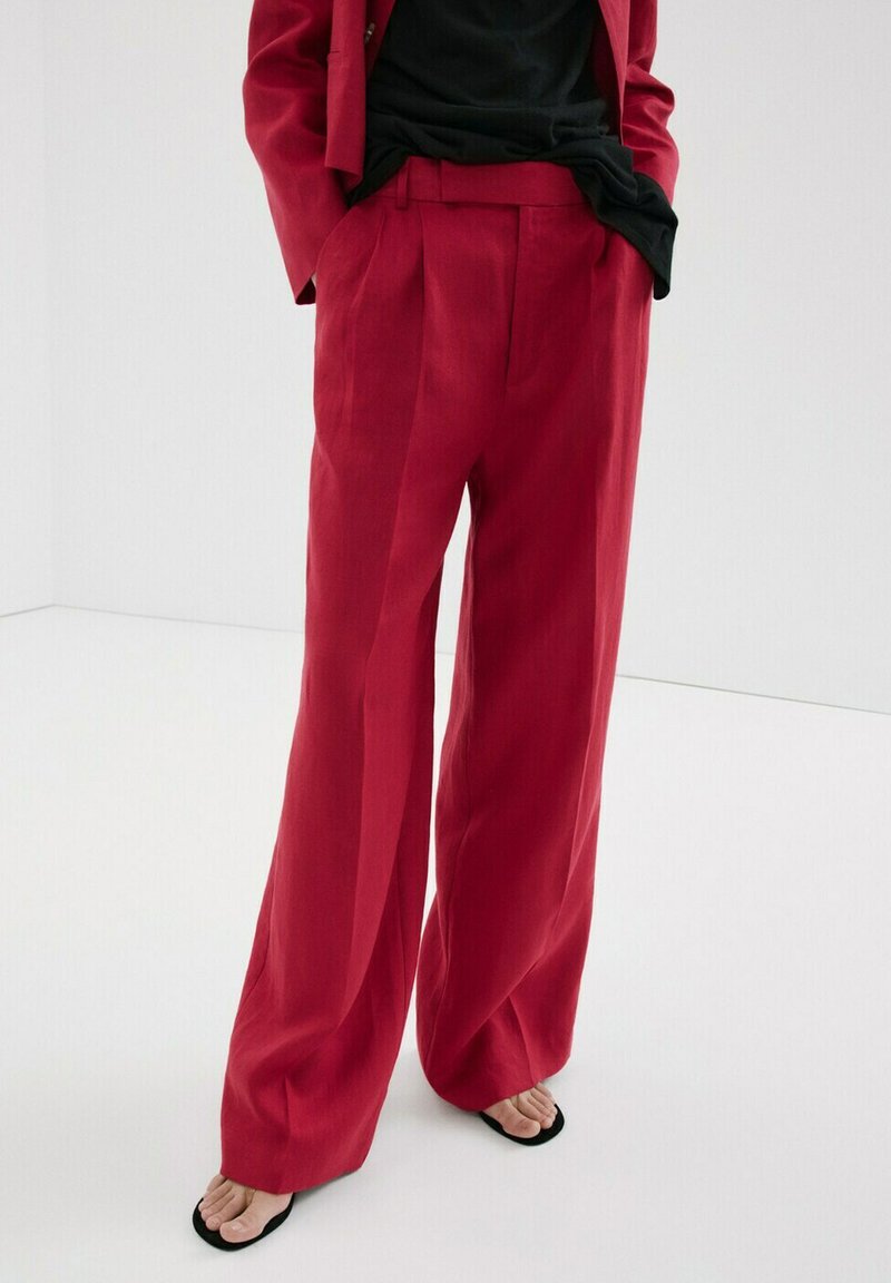 Persoon draagt een rood broek met hoge taille en wijde pijpen, handen in de zakken, gecombineerd met zwarte slippers en een deels zichtbaar zwart shirt.