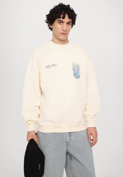 Cremefarbener Sweatshirt mit gerippten Bündchen, auf der Vorderseite mit einem blauen Grafikdruck und Text versehen. Getragen mit hellblauen Jeans und hält eine schwarze Tasche.