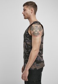 Tricou tip tank top cu model camuflaj în nuanțe închise, având un gât rotund și un design fără mâneci. Fabricat din bumbac, cu detalii vizibile ale unui tatuaj pe braț.