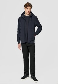 Mężczyzna w czarnej pikowanej kurtce z kapturem, czarnych spodniach i czarnych tenisówkach z białymi podeszwami, stojący z jedną ręką w kieszeni.