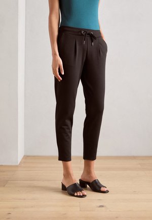 Black tapered pants with a drawstring waist, pleats, and side pockets, paired with black slide sandals on a wooden floor.