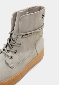 Bottine en suède gris avec un col rembourré, un bout rond et une semelle en caoutchouc plate. Dotée d'œillets en métal et de lacets gris assortis.