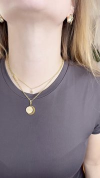 Collane d'oro stratificate intorno al collo con un pendente circolare contrassegnato "GUESS", indossate su una t-shirt aderente grigia scura.