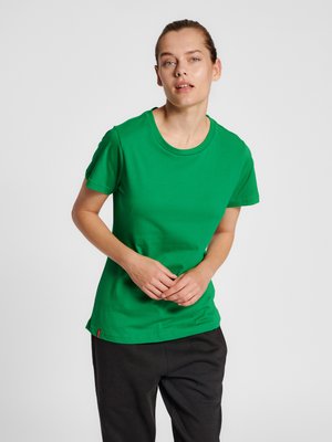 Femme à la peau claire et aux cheveux attachés, portant un t-shirt vert à manches courtes et un pantalon noir, debout devant un fond gris clair uni.