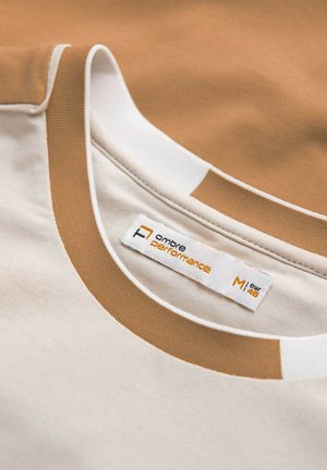 Camiseta cómoda con un cuerpo beige claro y acentos marrones. Presenta un escote redondo con ribete blanco y una etiqueta que indica la talla y la marca.