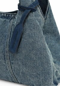 Sac à bandoulière en denim bleu foncé, présentant une texture douce, une forme incurvée, et une étiquette attachée à la sangle avec un texte en relief.