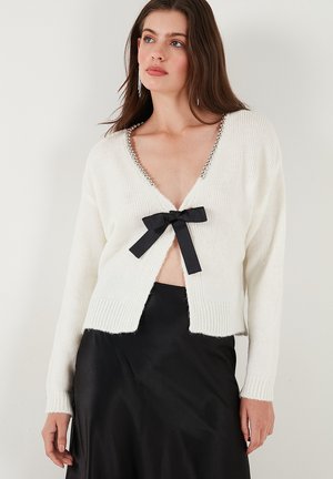 White knitted cardigan featuring a deep V-neck, embellished neckline with silver accents, and a black bow at the front. Soft texture.