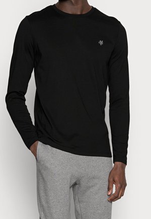 Homme portant une chemise noire à manches longues avec un petit logo sur la poitrine et un pantalon gris clair, debout avec une main dans la poche.