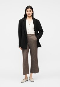 Femme debout, main dans la poche, portant un blazer noir, un haut blanc boutonné, un pantalon large marron et des chaussures beige à enfiler.