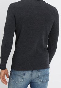 Ciemnoszary sweter dzianinowy z długimi rękawami, teksturowany materiał, prążkowany kołnierz i subtelny detal z tyłu, zestawiony z jasnoniebieskimi dżinsami.