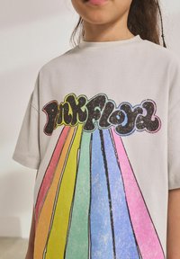 Svetlosivá bavlnená tričko s nápisom "Pink Floyd" v čiernej farbe, z ktorej vychádzajú dúhové pruhy, textúrovaný dizajn.