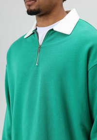 Groene trui met een witte, gestructureerde kraag en een halve rits aan de voorkant. De stof lijkt glad en zacht, wat een casual uitstraling geeft.