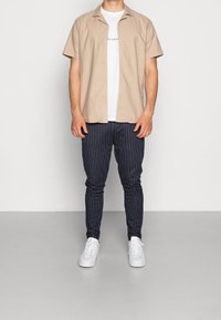 Beige short-sleeve button-up shirt over a white t-shirt, paired with navy striped jogger pants and white sneakers.