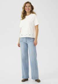 Weißes T-Shirt mit gewelltem, besticktem Saum, kurzen Ärmeln, kombiniert mit hellblauen, weiten Jeans und braunen Slippers.