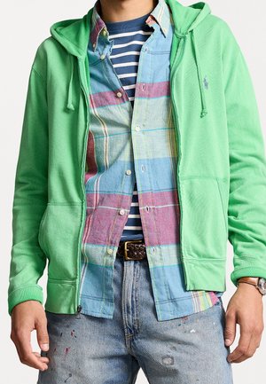 Homme portant un sweat à capuche vert zippé par-dessus une chemise à carreaux bleue, rose et blanche boutonnée, ainsi qu'un t-shirt rayé, avec un jean éclaboussé de peinture et une ceinture marron.