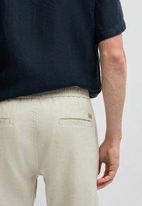 Light beige trousers with a relaxed fit, featuring side pockets and a small logo patch. Paired with a dark, textured short-sleeve shirt.