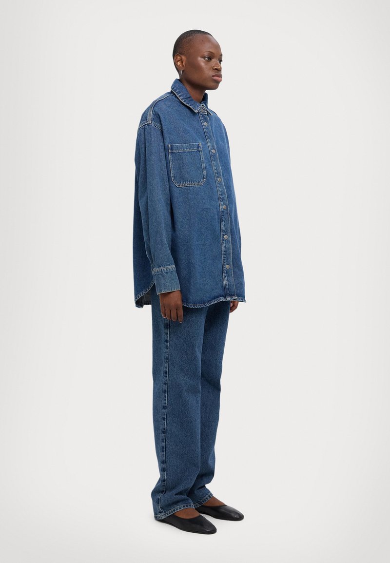 Overdimensjonert denimskjorte med krage, frontlomme og knappelukking, assortert med løstsittende denimbukser, alt i en mellomblå nyanse.