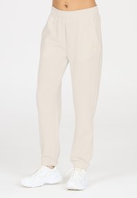 Hellbeige Sweatpants mit elastischem Bund, Seitentaschen und gerippten Bündchen, kombiniert mit weißen Sneakern. Glatte, weiche Textur.
