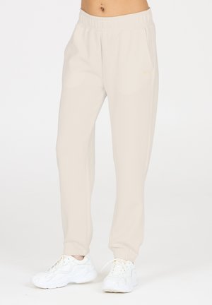 Hellbeige Sweatpants mit elastischem Bund, Seitentaschen und gerippten Bündchen, kombiniert mit weißen Sneakern. Glatte, weiche Textur.