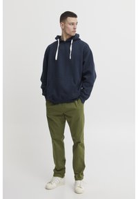 Tmavo modrá mikina s kapucňou a bielymi šnúrkami, klokaní vrecko, spárovaná s olivovozelenými nohavicami a bielymi teniskami.