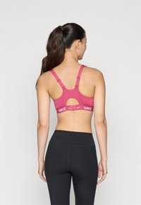 Rózsaszín sportmelltartó kivágott hátú dizájnnal és lapos pántokkal. Fehér logószalaggal rendelkezik, rajta a 'NIKE' felirattal. Fekete, magasított derekú leggingszel párosítva.