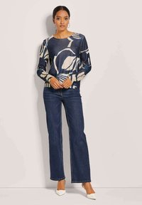 Langärmliger Strickoberteil mit einem blauen und beigen abstrakten Muster, kombiniert mit dunklen Jeans im Schlagstil und weißen High Heels.