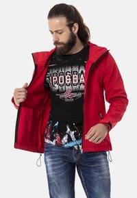 Rote wasserdichte Jacke mit Kapuze, Reißverschluss vorne und Taschen, über einem schwarzen Grafik-T-Shirt mit bunten Spritzmustern und Text.