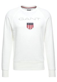 Sudadera blanca con mangas raglán, que presenta el logo "GANT" en gris y un parche en forma de escudo con diseño en rojo, blanco y azul.