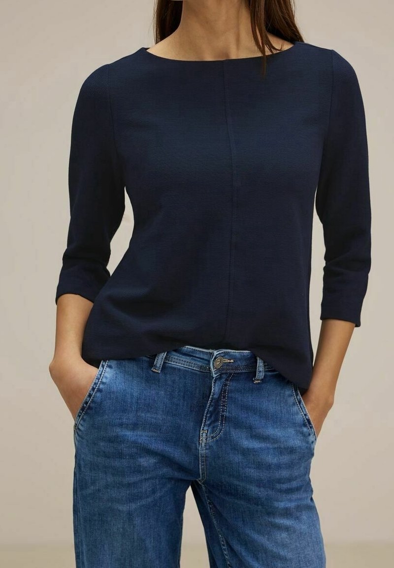 Námořnická modrá strukturovaná blůza s kulatým výstřihem a tříčtvrtečními rukávy, zkombinovan�á se světle modrými džíny z denimu s knoflíkem a kapsami.