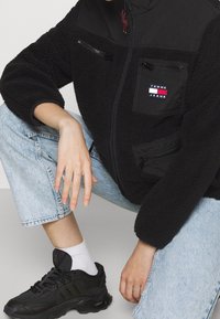 Czarna polarowa kurtka z zamykanymi na zamek kieszeniami, wysokim kołnierzem i kontrastującymi ramionami. Noszona z jasnoniebieskimi dżinsami i czarnymi trampkami.