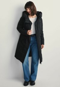 Kobieta w długim czarnym ocieplanym płaszczu z kapturem, białej bluzce, niebieskich dżinsach i czarnych botkach, stojąca przy gładkiej jasnej ścianie.