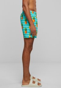 Homme portant un maillot de bain turquoise avec des ananas oranges et un cordon blanc, associé à des sandales en toile blanc cassé, se tenant de profil.