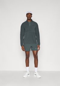 Pullover in pile verde scuro con zip a quarto, abbinato a pantaloni corti coordinati. Sneakers bianche e un cappellino bianco completano l'outfit.