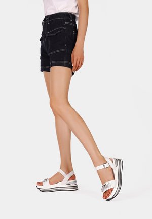 Shorts di denim nero con tasche frontali, cuciture bianche e una forma aderente, abbinati a sandali bianchi con un accento argentato lucido e suola a strati.