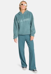 Teal, fleece pullover met capuchon en halve rits, met de tekst "BILLABONG"; gecombineerd met rib gebreide teal broek en zwarte sneakers.