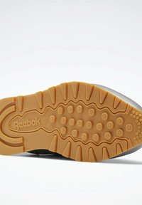 Musta tennaripohja, jossa on teksturoitu, hunajankeltainen kumipohja, jossa on pyöreitä nystyröitä ja uurteisia kuvioita; "Reebok"-logo on painettu.