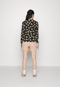 Blusa nera a fiori con fiori beige, maniche lunghe e dettagli a fiocco. Abbinata a pantaloni corti rosa chiaro e sneakers beige, mostrata da dietro.
