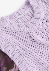 Pull en laine lavande avec un motif texturé et des bords côtes, arborant une étiquette de la marque "NEXT." Superposé sur un tissu à imprimé floral.