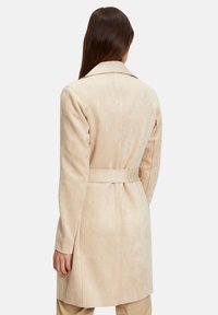 Beige pitkä takki, jossa on sileä, mokkamainen pinta. Siinä on vyötäröllä solmittava vyö, leveät kaulukset ja suoraviivainen leikkaus.