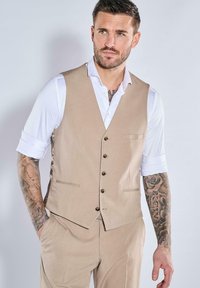 Beige maßgeschneiderte Weste mit Reverskragen, Fünf-Knopf-Verschluss und Taschen, getragen über einem weißen Hemd. Verfügt über eine glatte Textur.