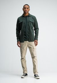 Grüne Button-up-Hemd, dunkelgrünes T-Shirt, beige Cargo-Hosen und schwarze Sneaker mit weißen Sohlen. Lässiges Outfit mit functionalem Design.
