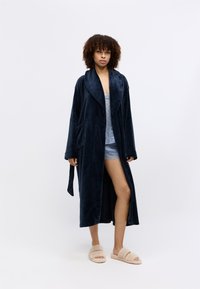 Tummansininen pörröinen aamutakki, jossa on kauluskotelo, sivutaskut ja nauhakiristys. Päällä vaaleansininen pitsireunainen toppi ja beigekulhoiset tohvelit.