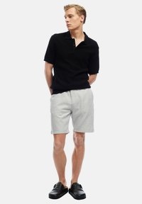 Jonge man staat met zijn handen achter zijn rug, gekleed in een zwart poloshirt met korte mouwen, lichtgrijze korte broek en zwarte instap schoenen, tegen een witte achtergrond.